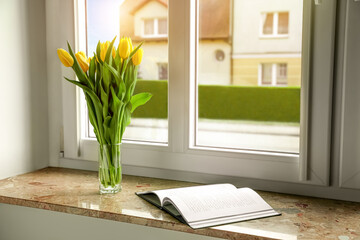 Bouquet of beautiful yellow tulip flowers in glass vase and open book on windowsill. Space for text
