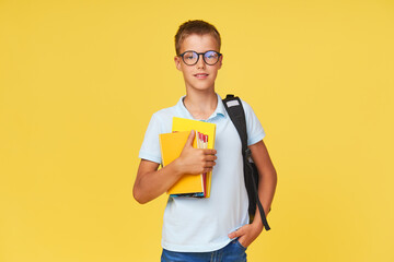 Portrait of a schoolboy in glasses with textbooks and a backpack on a yellow background. Back to...