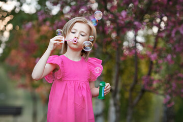Child girl in pink dress blowing soap bubbles in nature against background of blooming tree
