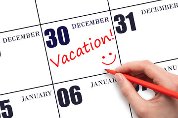 A hand writing a VACATION text and drawing a smiling face on a calendar date 30 December. Vacation planning concept.