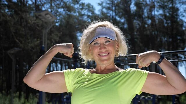Mature woman shows powerful arm biceps against green park. Middle-aged blonde lady wearing white cap sun visor enjoys healthy life on sports ground