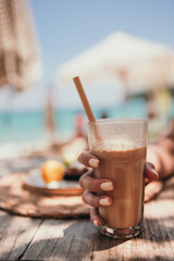 Woman hand with yellow manicure hold ice coffee latte in tall glass with straw by the sea in beach bar