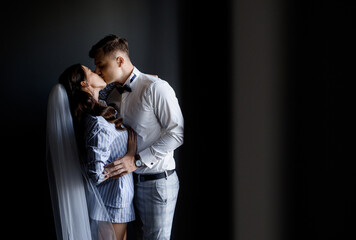 Handsome groom man kissing bride in pajama and veil in curly hairstyle and hugging. Loving brides couple enjoying each other, waiting for wedding ceremony.