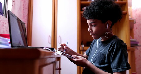 Mixed race kids doing homework at home writing on notepad and looking at laptop screen