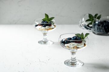 Two glasses with a healthy breakfast. Granola, Greek yogurt, blueberries, mint. In the background is a jar of blueberries. Light background.