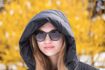 portrait of a young girl with long hair, wearing dark glasses, in a black jacket with a hood put on her head, against the background of yellow foliage