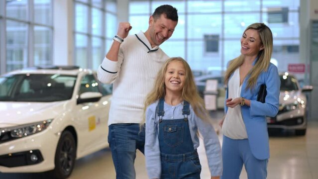 Cheerful cute girl dancing in car dealership looking at camera as blurred parents smiling talking at background. Portrait of pretty joyful Caucasian child rejoicing new automobile purchase in showroom