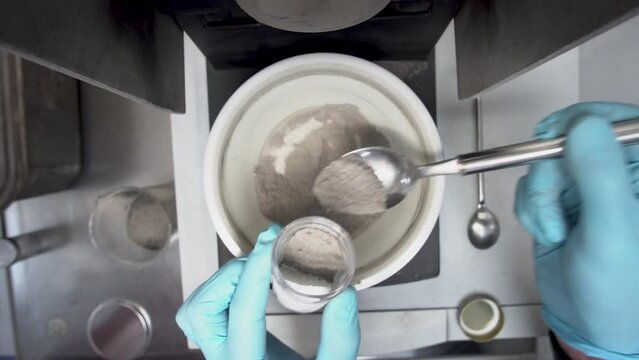 Lab worker scoops powder from mill into a container for testing, overhead