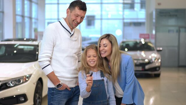 Portrait of happy pretty girl posing with man and woman in car dealership bragging key. Cute Caucasian daughter rejoicing new vehicle purchase with father and mother indoors. Excitement and richness