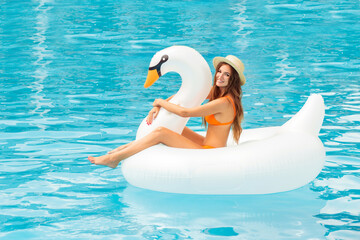 A sexy girl in an orange swimsuit lies on an inflatable mattress in the pool. Woman smiling in bikini on floating white swan.