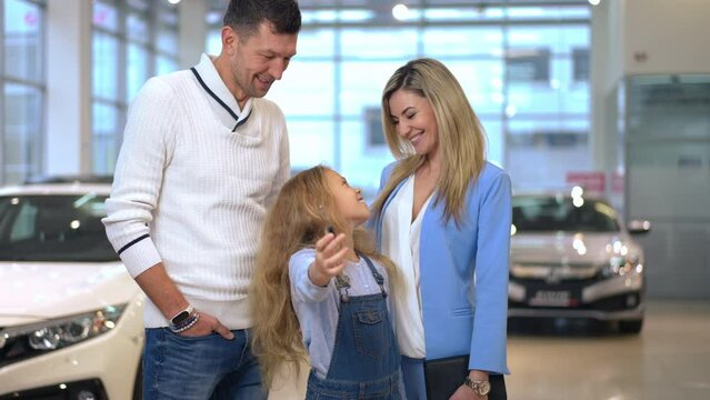 Live camera zoom in to happy girl showing car key smiling standing with mother and father in car dealership. Excited Caucasian daughter enjoying buying new vehicle in showroom with parents