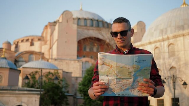 Man tourist looking at paper map against Ayasofya (Hagia Sophia) Mosque, travel and sightseeing. Guy wearing shirt holding and navigating with topographic map by city, popular area in Istanbul Fatih