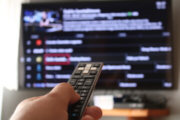 Hand with a remote control, in the background is a TV with a list of channels.