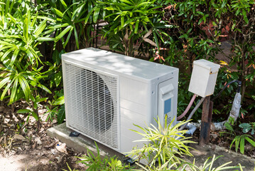 A split type inverter air conditioning condenser mounted outside a house partially concealed with shrubs. HVAC for tropical countries. - obrazy, fototapety, plakaty