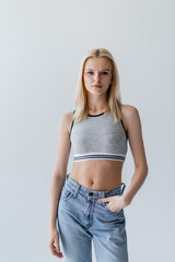 Pretty blonde woman in jeans and top standing isolated on grey.