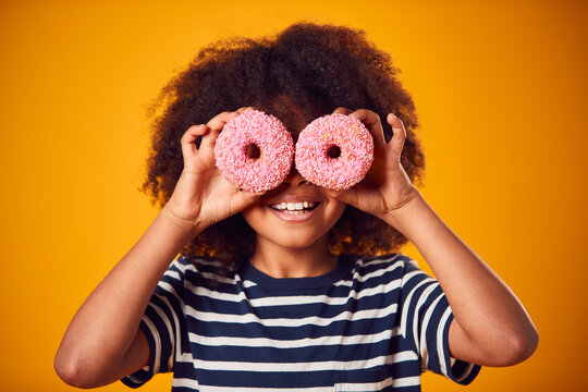 Studio Portrait Of Boy Holding Two Donuts In Front Of Eyes Against Yellow Background