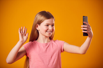 Studio Portrait Of Young Girl Posing For Selfie On Mobile Phone Against Yellow Background