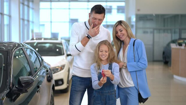 Portrait of cute Caucasian girl playing with toy car choosing new automobile with parents in dealership. Pretty daughter beautiful mother and handsome father talking smiling buying vehicle in showroom