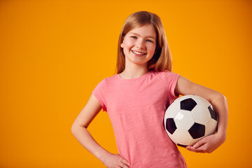 Studio Shot Of Young Girl Holding Soccer Ball Under Arm Against Yellow Background