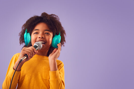 Studio Shot Of Boy Wearing Headphones Singing Karaoke Into Microphone Against Purple Background