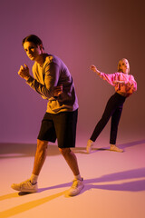 Full length of stylish couple in sweatshirts and sneakers on purple background with lighting.