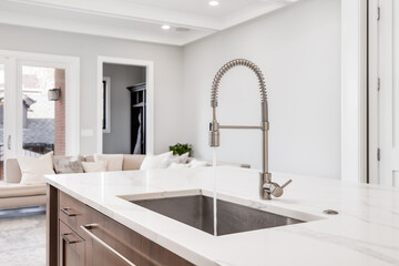 Fototapeta na wymiar A kitchen sink with water running in a luxurious home looking out towards a living room area. The kitchen island features a white granite and stainless steel tub.