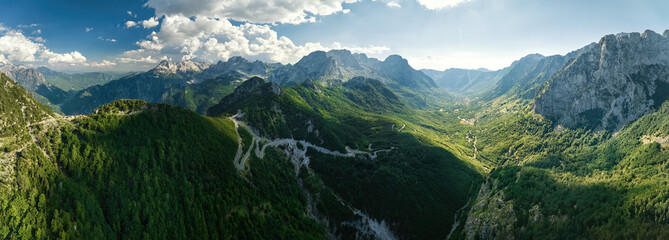 A panoramic, aerial view of the winding road into the Theth Valley. Green valleys, blue sky with clouds, steep rocks, remnants of snow. Theth national park, Albanian Alps, Albania.