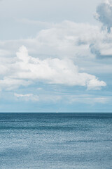 A single sailing boat on the horizon in the centre of the frame. Blue waves and ocean in foreground with fluffy white clouds above. Copy space available
