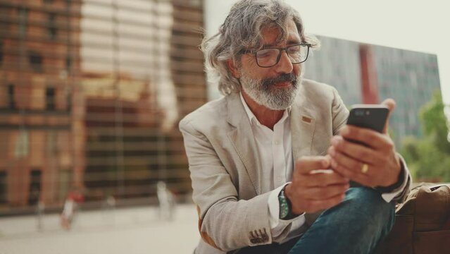 Mature businessman with beard in eyeglasses wearing gray jacket is using cell phone. Middle aged manager scrolling information on his smartphone while sitting outside the office