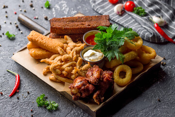 assorted beer snacks, chicken wings, fried croutons, onion rings, cheese sticks and chicken popcorn on board with sauce on grey table