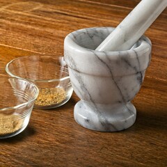 Close-up of marble mortar and pestle and prep bowls of ground spices on a wooden tabletop side lit by sunny window