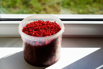 .a big beautiful transparent bucket of red currants with berries on a white windowsill on a sunny day