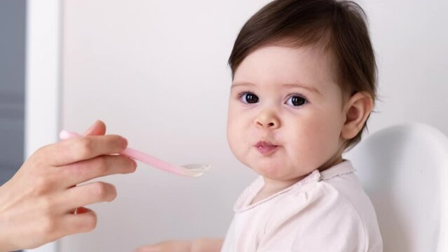 Baby girl eating blend mashed food sitting, on high chair, mother feeding child, hand with spoon for vegetable lunch, baby weaning, first solid food for young kid.