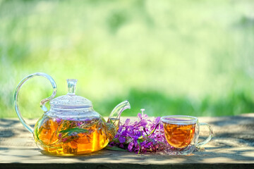 Glass teapot and cup with tea from flowers of fireweed on wooden table in garden, abstract green...
