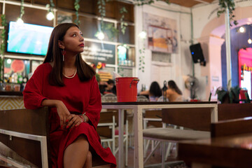 Brunette Latin woman dressed in red sitting at a bar table waiting for her waiter. Hispanic girl in a nightclub looking to the side. Party girl in a bar with a red outfit.