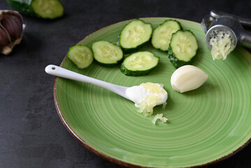 squeezed garlic and fresh cucumbers cut into slices on a green plate. white spoon of sour cream.