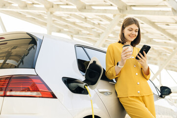 Vehicle charging at public charging station near solar power plant. Woman with a mobile phone and...