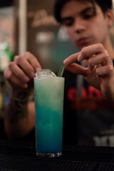 Defocused bartender preparing a blue colorful drink in a nightclub. A blue alcoholic drink with ice and a leaf topper. Waiter preparing a drink for a client. Drinks in nightclubs.
