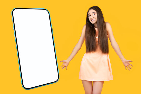Positive puzzled asian girl with arms to the sides and a funny smile on a yellow background near the mockup cellphone with white blank screen.