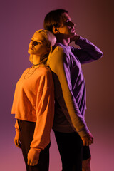 Stylish couple standing back to back on purple background with lighting.