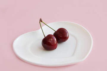 two cherries on a sprig on a white plate in the form of a heart on a light background