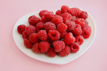 large ripe raspberries in a white plate in the shape of a heart on a pink background