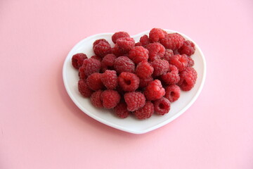 large ripe raspberries in a white plate in the shape of a heart on a pink background