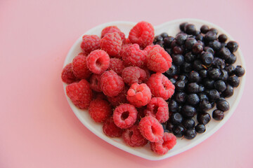 ripe raspberries and blueberries on a white plate in the shape of a heart on a pink background
