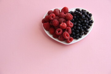 ripe raspberries and blueberries on a white plate in the shape of a heart on a light background close-up