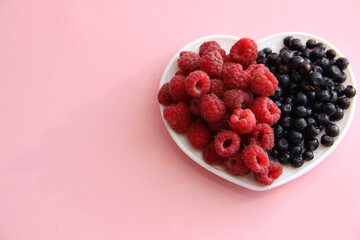 Large raspberries and blueberries on a white plate in the form of a heart on a pink background