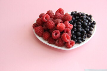 ripe raspberries and blueberries on a white plate in the shape of a heart on a light background close-up