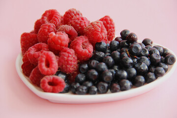 ripe raspberries and blueberries on a white plate in the shape of a heart on a light background close-up