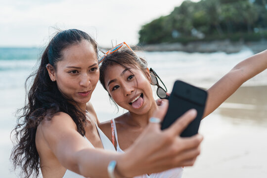 Portrait of happy smiling beautiful women taking a selfie photo with mobile phone on the beach. Pretty young Asian girls in swimwear posing and for picture together on tropical vacation