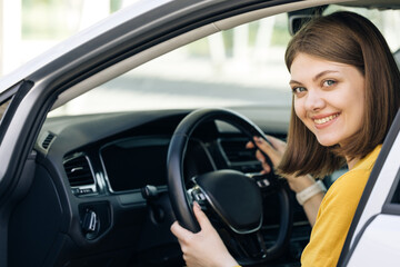Fototapeta na wymiar Attractive young business woman looking over her shoulder while driving a car. Driver smiles at camera. Happy millennial woman taxi driver sitting in car holding to steering wheel.